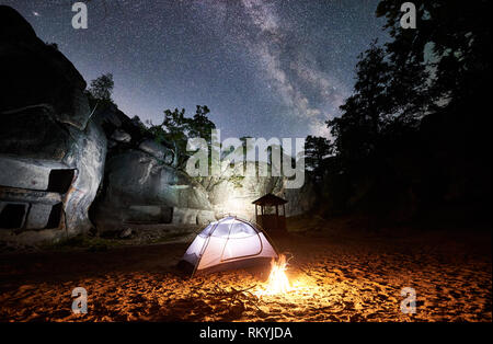 Camping im Sommer Nacht in der Nähe von Felsen. Weißglühend touristische Zelt und Lagerfeuer unter erstaunlichen Nacht Himmel voller Sterne und Milchstraße. Auf dem Hintergrund schöner Sternenhimmel, große Steine und Bäume Stockfoto