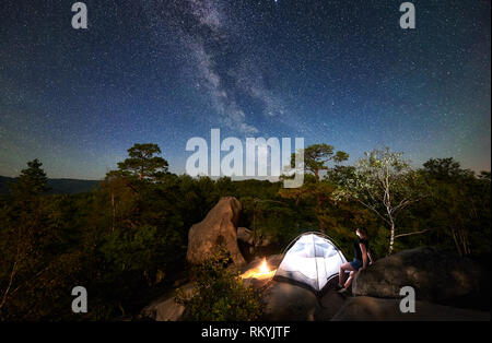 Frau Wanderer ruht auf Rocky Mountain Top neben Camp, Bonfire und touristische Hütte im Sommer Nacht, mit Sicht auf den Himmel voller Sterne und Milchstraße. Auf Hintergrund Sternenhimmel, große Steine und Bäume Stockfoto