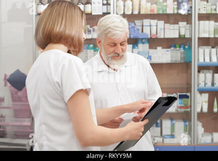 Älterer Mann, Beratung mit weiblichen Apotheker in der Apotheke. Frau in Weiß einheitliche Holding schwarzen Ordner, helfen, die Infos über Medikamente. Alter Mann mit grauen Haaren lächelnd. Stockfoto