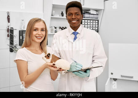 Schönen afrikanischen Tierarzt, Ordner, in der Tierklinik mit dem Besitzer von Hamster posieren. Wunderschöne und charmante Frau mit Pet in den Händen. Frau und Fachmann an der Kamera schauen und lächeln. Stockfoto