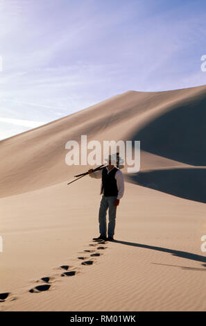 Porträt eines lächelnden Mitte erwachsenen Mann hält Fotografie Ausrüstung beim stehen vor einer großen sanddüne in der Wüste. Stockfoto