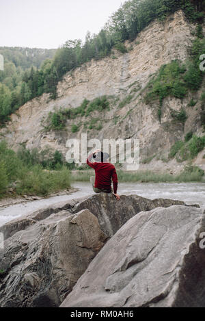 Junge Mann in roter Jacke und schwarzen Hut steht auf einem Felsen und genießt. Epische Berge im Hintergrund. Concept outdoor Natur lifestyle Stockfoto