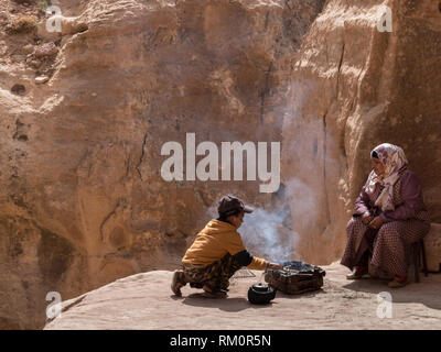 Ein junger Beduine junge bereitet sich sorgfältig ein dampfendes warmes Essen und Kaffee unter den Augen seiner Mutter an der Basis von einem entfernten Höhle in Petra in Jordanien. Stockfoto