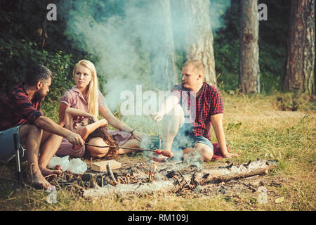 Gruppe von Freunden camping im Wald. Menschen Spaß sprechen um Lagerfeuer. Kochen Würstchen über dem Feuer Stockfoto