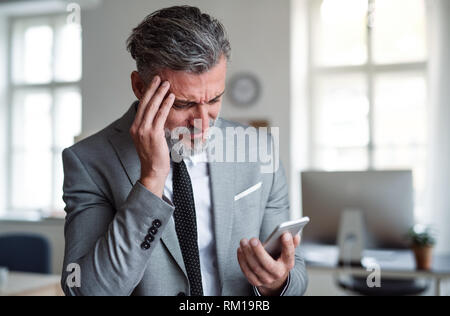 Ein frustrierter Geschäftsmann mit Smartphone steht in einem Büro, lesen schlechte Nachrichten. Stockfoto