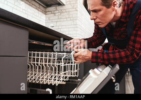 Reparatur Geschirrspüler. Männliche Techniker in der Nähe von Spülmaschine sitzen mit Schraubendreher in Küche mit Instrumenten Stockfoto