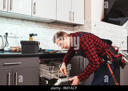 Alle Instrumente sind im Preis inbegriffen. Männliche Techniker stehen in der Nähe von Spülmaschine mit Schraubendreher in Küche mit Instrumenten Stockfoto