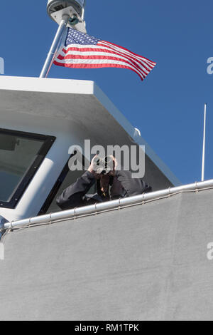 Mann schaut durch ein Fernglas von der Brücke eines Whale Watching Boot mit der amerikanischen Sterne und Streifen der Flagge über ihm. Stockfoto