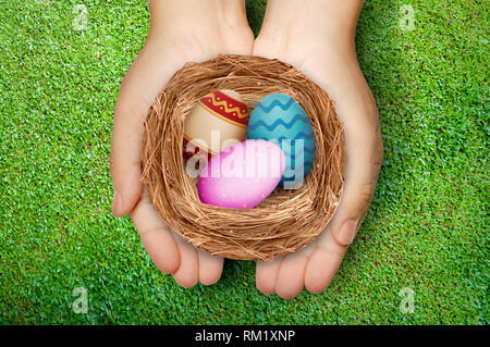 Menschliche Hände halten Nest mit bunten Ostereiern über grüne Gras Hintergrund. Frohe Ostern Stockfoto