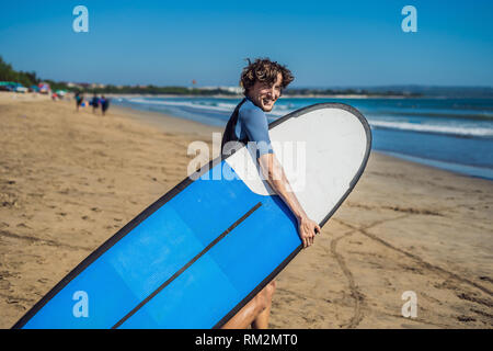 Schöne sportliche junge Surfer mit seinem Surfboard unter dem Arm in seinem Anzug posiert an einem Sandstrand der tropischen Strand Stockfoto