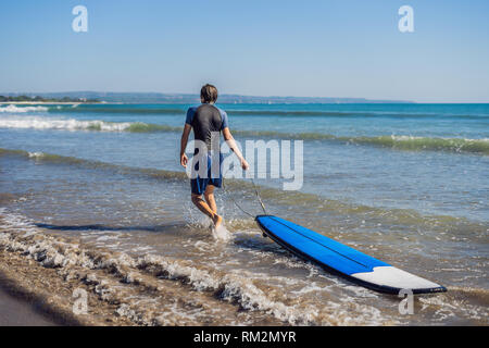 Schöne sportliche junge Surfer mit seinem Surfboard unter dem Arm in seinem Anzug posiert an einem Sandstrand der tropischen Strand Stockfoto