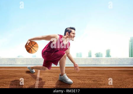 Junge asiatischer Mann Basketballspieler im Dribbling den Ball zwischen den Beinen auf Baskettballplatz draußen mit Stadtansichten und blauer Himmel Hintergrund posiert Stockfoto