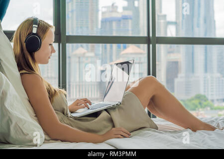 Junge Frau, die an einem Notebook arbeitet in seinem Bett auf einem Hintergrund von einem Panoramafenster mit Blick auf die Wolkenkratzer. Nutzt drahtlose Kopfhörer. Freiberufler Stockfoto