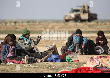 Deir Ezzor, Syrien. 12 Feb, 2019. Eine Familie wartet mit ihrem Gepäck backdropped von einem gepanzerten Fahrzeug als Zivilisten der islamische Staat fliehen - statt Dorf Baghouz im östlichen syrischen Provinz von Deir Ezzor. Mehr als 1.500 Menschen haben im vergangenen pocket Islamischen Staat im östlichen Syrien in den letzten 48 Stunden Links zu fliehen, die Schlacht zwischen den Kämpfern der syrischen Demokratischen Kräfte (SDF), einer von den USA unterstützten Kurdischen-Gruppe führte, und der Islamischen Staat. Credit: Aboud Hamam/dpa/Alamy leben Nachrichten Stockfoto