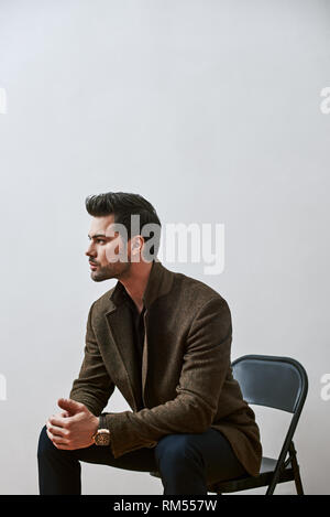 Portrait einer jungen gutaussehenden Mann, Braune Jacke, auf einem Stuhl sitzend mit den Händen auf seine Knie, Suchen auf weißem Hintergrund Stockfoto