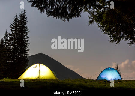 Zwei hell leuchtende, touristische Zelte auf der grünen Wiese Wald Lichtung auf dunklen Berg und dem klaren, blauen Sternenhimmel Kopie Raum Hintergrund. Tourismus, Nacht campin Stockfoto