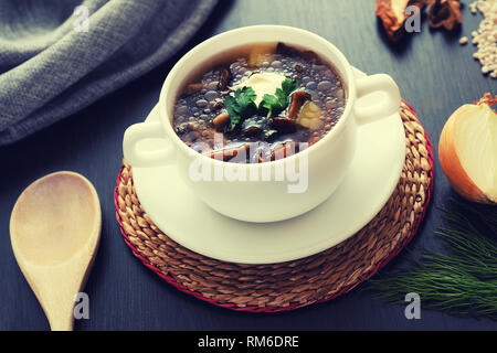 Zusammensetzung mit Pilz Suppe in einer Tasse mit Henkel, frische und getrocknete Pilze, auf einem Holztisch, auf einem Hintergrund von sackleinen. Löffel aus Holz. Dill und Zwiebeln. Stockfoto