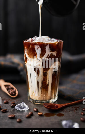 Eis Kaffee in ein hohes Glas mit Sahne über und Kaffeebohnen auf dunklem Beton Tabelle über schwarzen Holz- Hintergrund gegossen. Kalten Sommer trinken. Platz kopieren Stockfoto