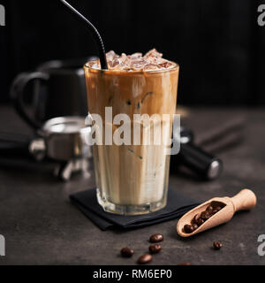 Eis Kaffee mit Sahne in ein hohes Glas und Kaffeebohnen, portafilter, Sabotage und kanne Milch auf dunklem Beton Tabelle über schwarzen Holz- Hintergrund. Kalte Summe Stockfoto