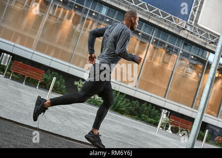 Gesunde Gewohnheiten. Volle Länge des Jungen und athletische afrikanischen Mann in stilvolle Sportswear laufen in die Stadt, während seine morgendlichen Training. Von der Seite. Gesundheit. Sport Konzept. Ausbildung Stockfoto