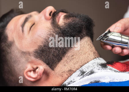Friseur Rasieren der Bart von einem stattlichen bärtiger Mann mit einem elektrischen Rasierer an der Friseur. Stockfoto