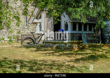 Jevington, Sussex, England, Großbritannien - 1 August, 2018: der Mann, der mit seinem Fahrrad im Schatten sitzen außerhalb der Halle von St. Andrews Kirche in Jevington. Die Stockfoto