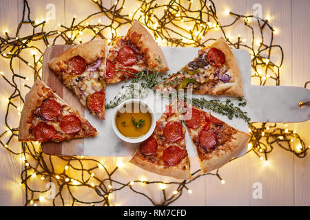 Weihnachten und Neujahr Atmosphäre. Heiße italienische Pizza mit Schmelzen mit Tomate, Salami und Käse auf einem weißen Marmor Schneidebrett. Hintergrund mit Beleuchtung Stockfoto