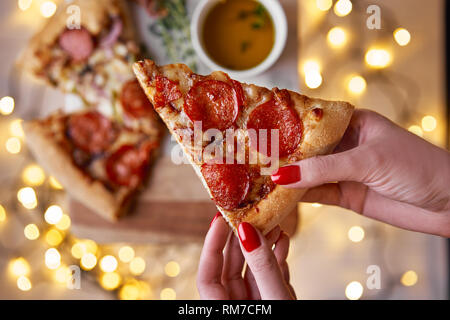 Weihnachten und Neujahr Atmosphäre. Womans hand nimmt Scheibe der Italienischen Pizza mit Schmelzen mit Tomate, Salami und Käse auf einem weißen Marmor Schneidebrett. Stockfoto