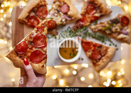 Weihnachten und Neujahr Atmosphäre. Womans hand nimmt Scheibe der Italienischen Pizza mit Schmelzen mit Tomate, Salami und Käse auf einem weißen Marmor Schneidebrett. Stockfoto