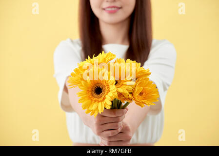 Schöne Frau im weißen Hemd mit Blumen Gerbera in den Händen auf einem gelben Hintergrund. Sie lächelt und lacht Stockfoto