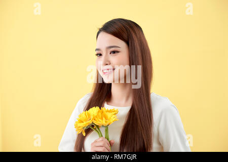 Schöne Frau im weißen Hemd mit Blumen Gerbera in den Händen auf einem gelben Hintergrund. Sie lächelt und lacht Stockfoto