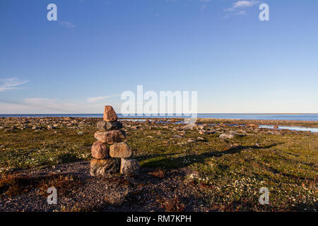 Inukshuk oder Inuksuk auf einem felsigen Tundra mit Wasser im Hintergrund Ende Juni in der Arktis Gemeinschaft der Arviat, Nunavut, Kanada Stockfoto