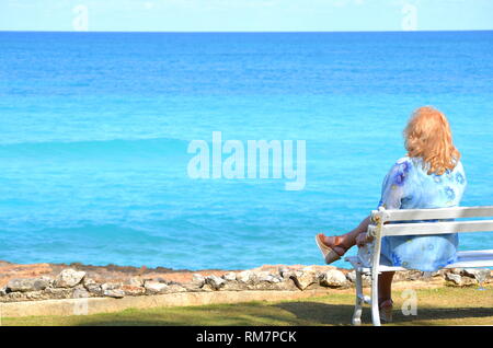 Senior Plus size blonde Frau sitzt auf der Bank mit Blick auf das blaue Meer mit kopieren. Solo Urlaub auf der tropischen Insel, Reisen, Freizeitaktivitäten und Self-care. Stockfoto