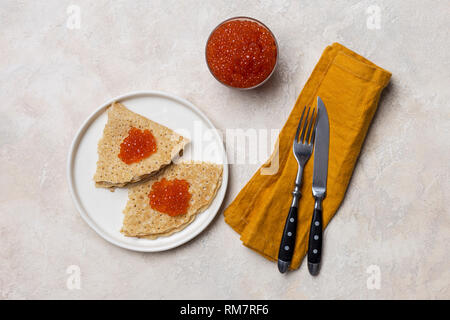 Frische thin Blini auf festliche mit rotem Kaviar, Gabel, Messer und Serviette auf weißem Hintergrund, Glas mit rotem Kaviar in der Nähe. Konzept der festlichen Essen. Sprach conce Stockfoto