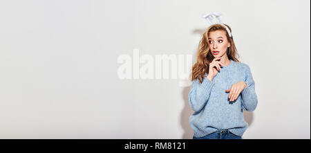 Portrait von verspielten nachdenkliches Mädchen mit blauen Pullover und Hasenohren die Hand am Kinn zur Seite mit geweitete Augen suchen, amazedly, auf weißem Hintergrund. Long Shot. Stockfoto