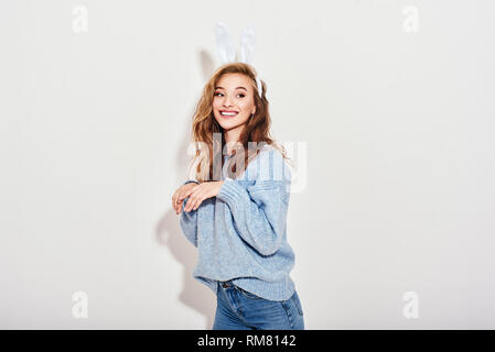 Portrait von verspielter, lebhafter Mädchen mit blauen Pullover, Jeans und Bunny Ohren, Hände wie Hase, interessiert beiseite suchen, auf weißem Hintergrund. Stockfoto