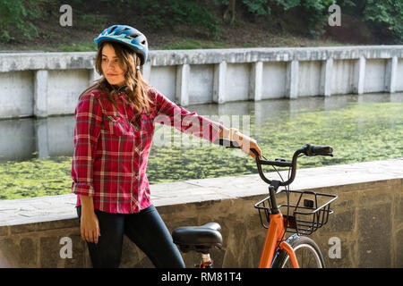 Fach ökologisches Verkehrsmittel Fahrrad. Schöne junge kasazy Frau trägt einen blauen Helm und lange Haare stellt neben eine orange-Farbe Stockfoto