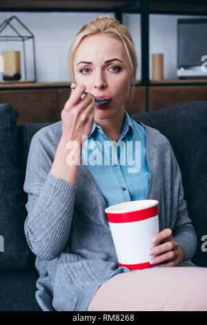 Traurige Frau mit Kamera suchen und essen Eis, während siitng auf der Couch zu Hause alleine Stockfoto