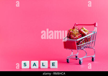 Klein Warenkorb mit Geschenken und Wort VERKAUF auf rosa Hintergrund. Rollwagen aus einem Supermarkt voller roter Geschenkboxen. Online shopping Konzept Schwarz F Stockfoto