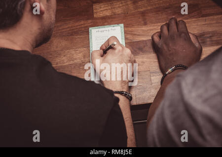 Ein interracial Gay Paare bilden eine Einkaufsliste für Lebensmittel auf einer hölzernen küche top zu Hause. Stockfoto