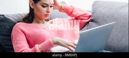 Attraktive indische Frau mit Bindi mit Laptop beim Liegen auf dem Sofa Stockfoto