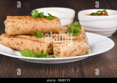 Pfannkuchen Brötchen mit Fleisch füllen, mit frischer Petersilie serviert, saure Sahne und Ketchup Sauce. Die traditionellen russischen Speisen auf Holz- Hintergrund. Nahaufnahme Stockfoto