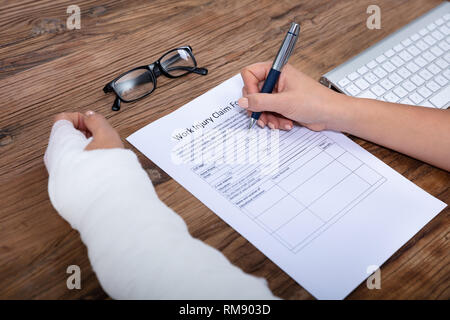 Nahaufnahme der Geschäftsfrau mit weißen Bandagen Hand Befüllen Arbeitsunfall Antragsformular auf hölzernen Schreibtisch Stockfoto