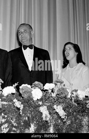 Präsident Lyndon B. Johnson steht aufmerksam, während eine junge Frau die Nationalhymne neben ihm bei einem Abendessen in das Conrad Hilton Hotel in Chicago singt, Ca.1964. Stockfoto