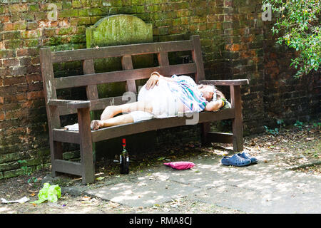 BIRMINGHAM, UK - März 2018 alte, betrunkene Frau Schlafen auf Holzbank. Flasche Alkohol, Tasche und Schuhe unter dem Sitz. Schmutzige Umgebung mit Gefallen Stockfoto