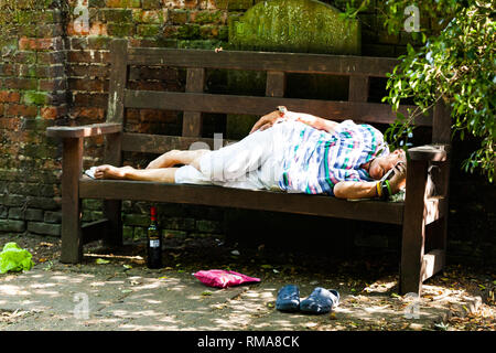 BIRMINGHAM, UK - März 2018 Ältere betrunkene Frau Schlafen auf Holzbank an einem sonnigen Tag. Flasche Fit Drink unter dem Sitz. Tasche und Schuhe auf Dir Stockfoto