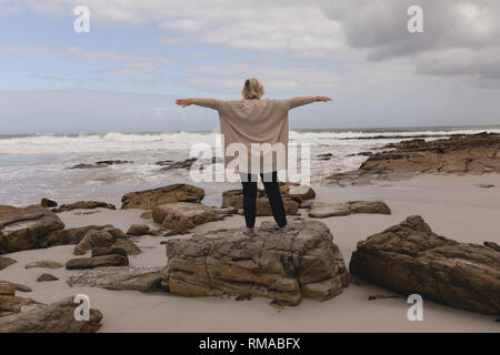 Ältere Frau, die mit den Armen auf den Felsen am Strand ausgestreckte Stockfoto