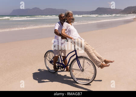 Älterer Mann reitet ein Fahrrad mit älteren Frau sitzt am Lenker am Strand Stockfoto