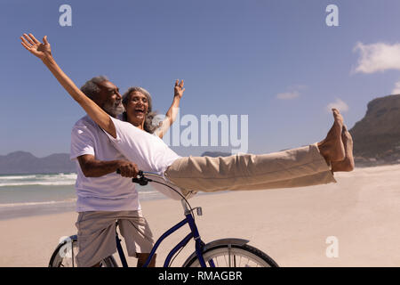 Älterer Mann reitet ein Fahrrad mit älteren Frau sitzt am Lenker am Strand Stockfoto