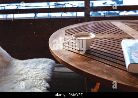 Holz-/Tabelle steht im Freien auf der Terrasse, lecker duftenden Cappuccino in einem weißen Schale, Dampf winkend. Buchen Sie in der Nähe der Schale liegen. Bank abgedeckt Stockfoto